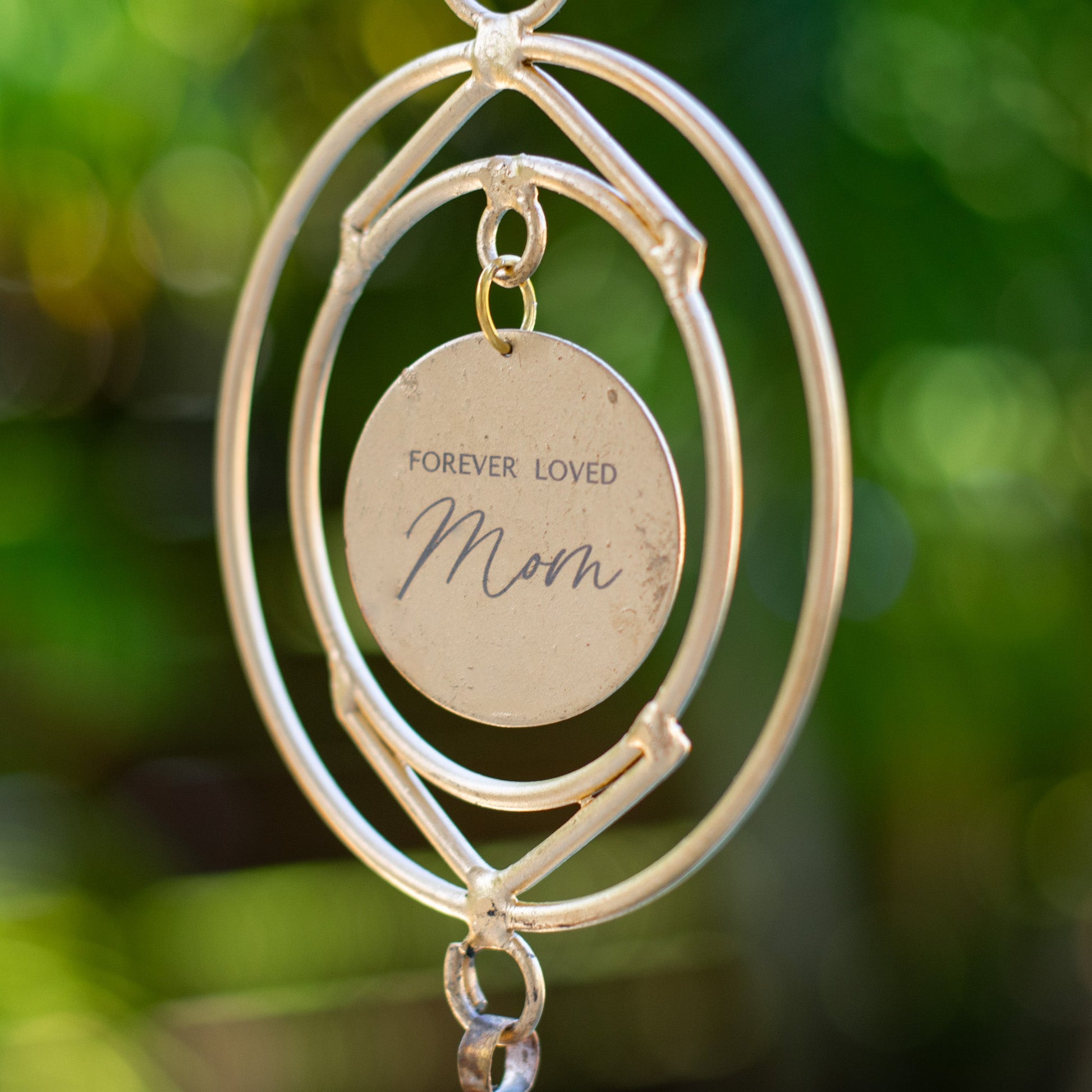 A close-up of a wind chime hanging outdoors, with a green garden background. The wind chime features two circular metal rings and a circular metal disk at the center with the engraved message: "FOREVER LOVED Mom". Below the disk, there is a small, rustic-style metal bell.