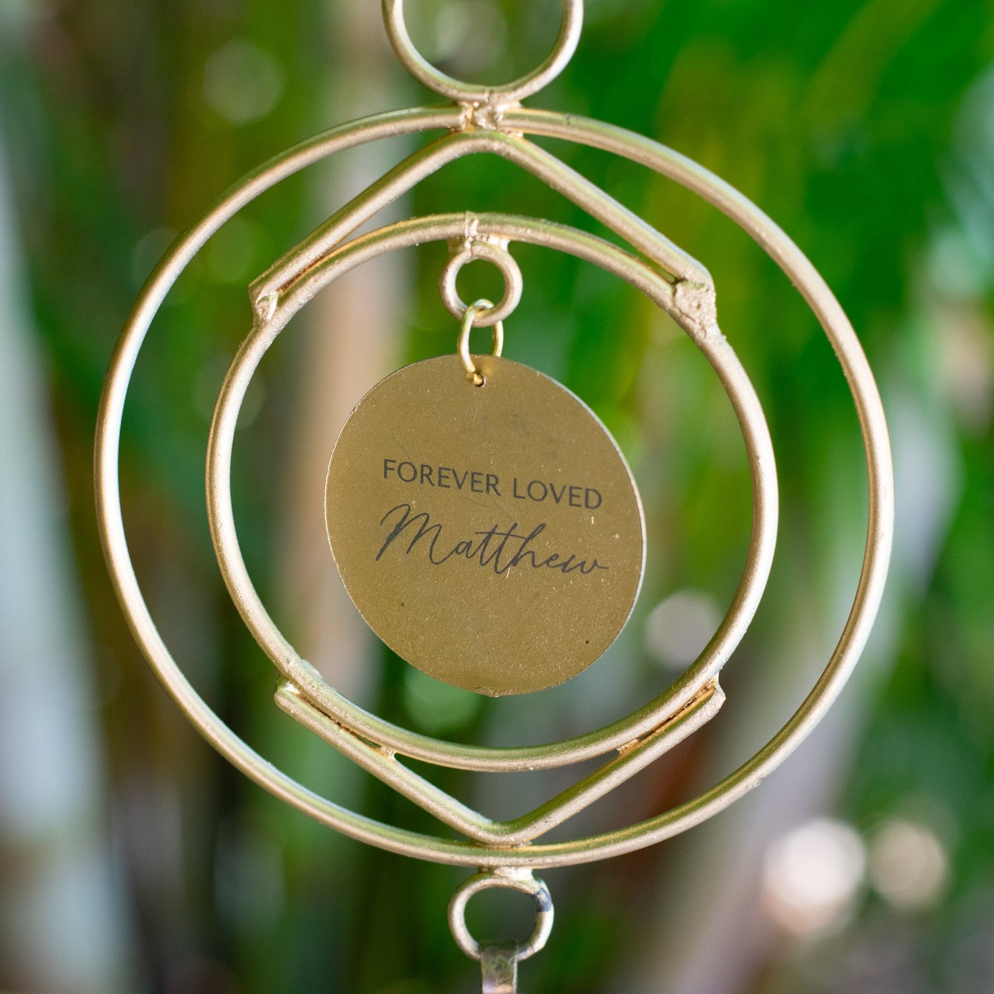 A close-up of a wind chime hanging outdoors, with a green garden background. The wind chime features two circular metal rings and a circular metal disk at the center with the engraved message: "FOREVER LOVED Matthew". Below the disk, there is a small, rustic-style metal bell.
