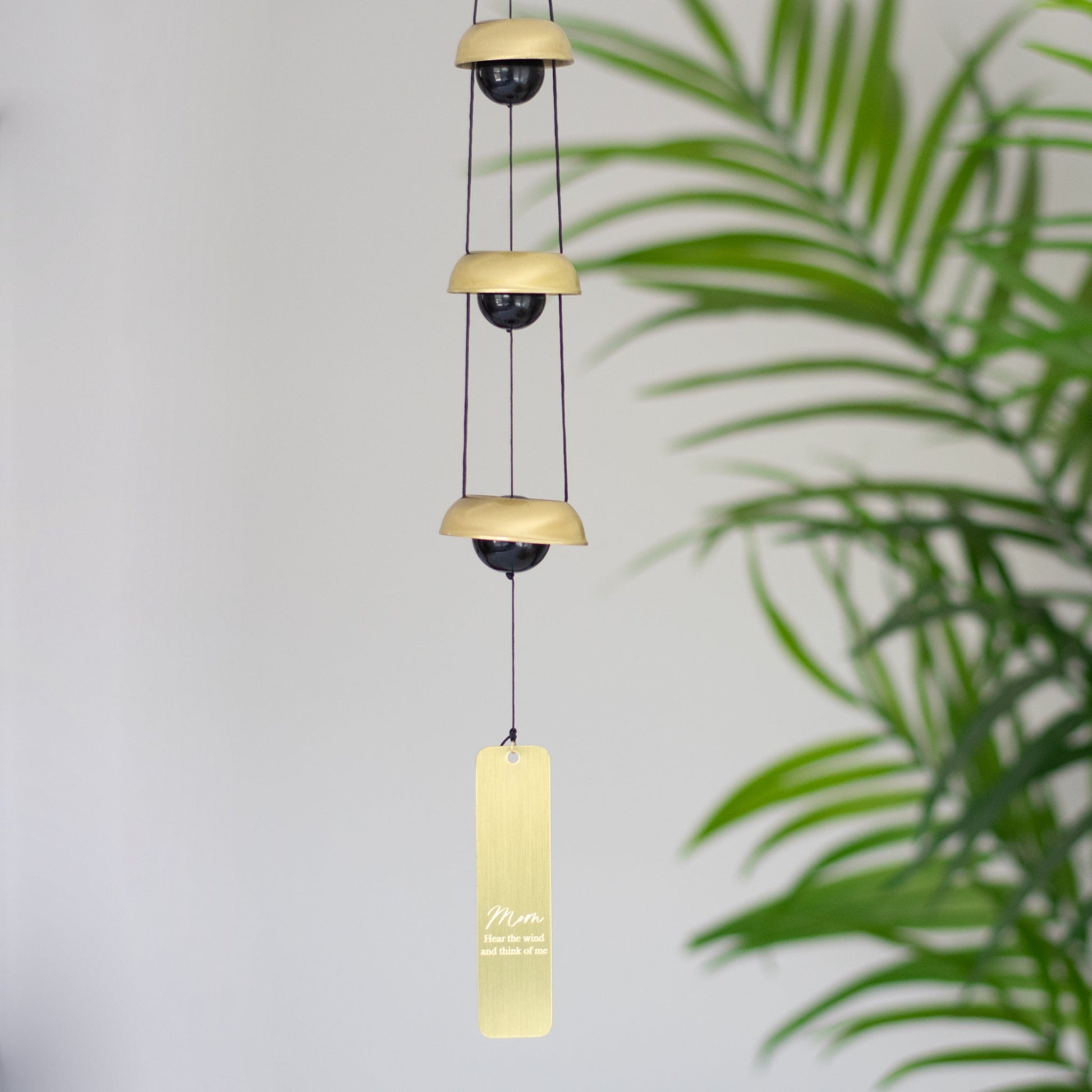 A wind chime hanging inside next to a plant that features three metallic bells, black cords, and a vertical rectangular tag at the bottom with the text mom and Hear the wind and think of me