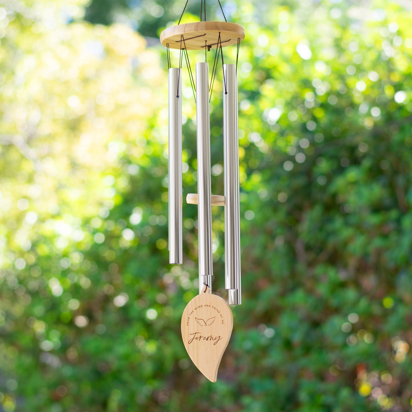 A wind chime handing outdoors features long metal tubes with a wooden disc at the top. A wooden leaf is at the bottom with the text Jeremy and Hear the wind and think of me