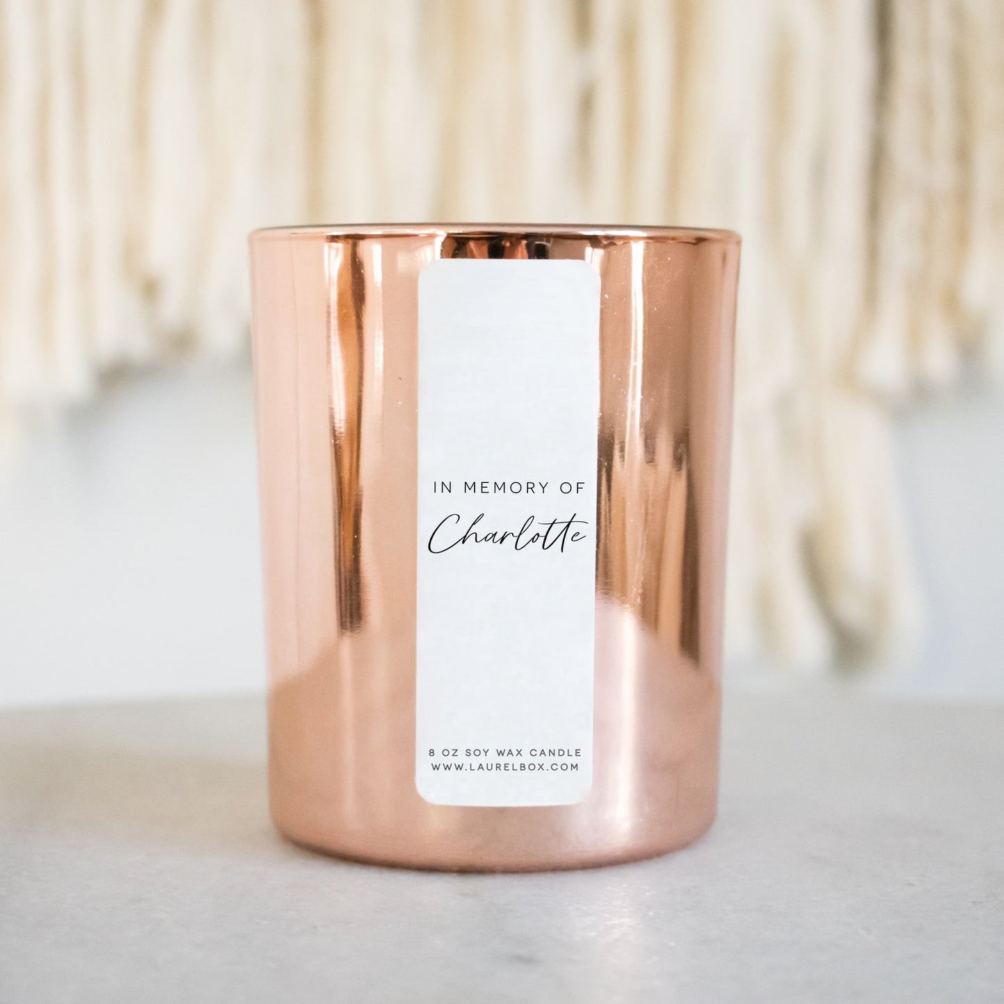 A gold-coated candle with a white sticker with the text In Memory of Charlotte sitting on a white desk